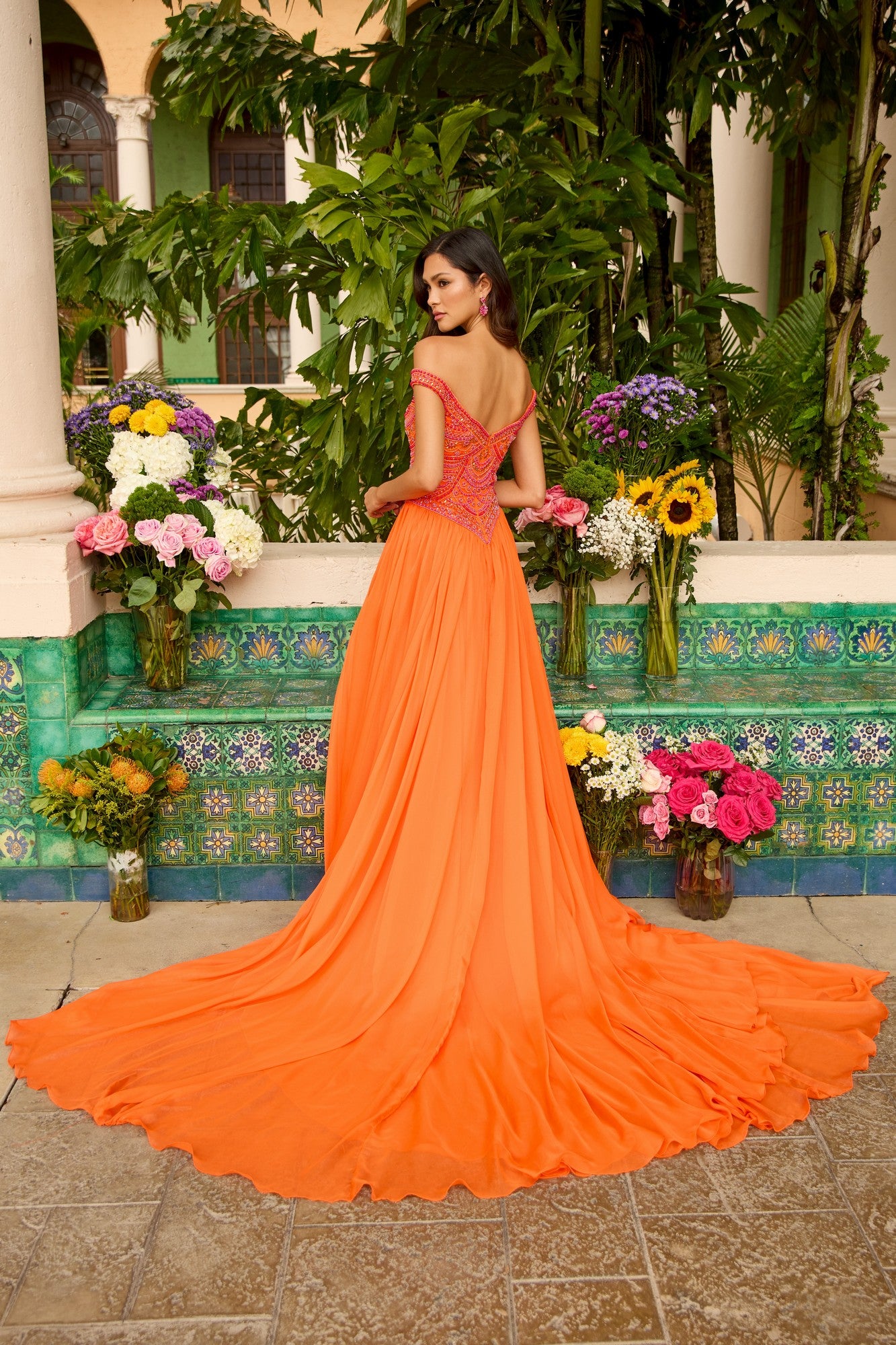 A woman stands with her back turned in an outdoor garden, wearing the Ava Presley 47254 long prom dress—an orange off-the-shoulder gown with a beaded bodice and long train, surrounded by colorful flowers and greenery.